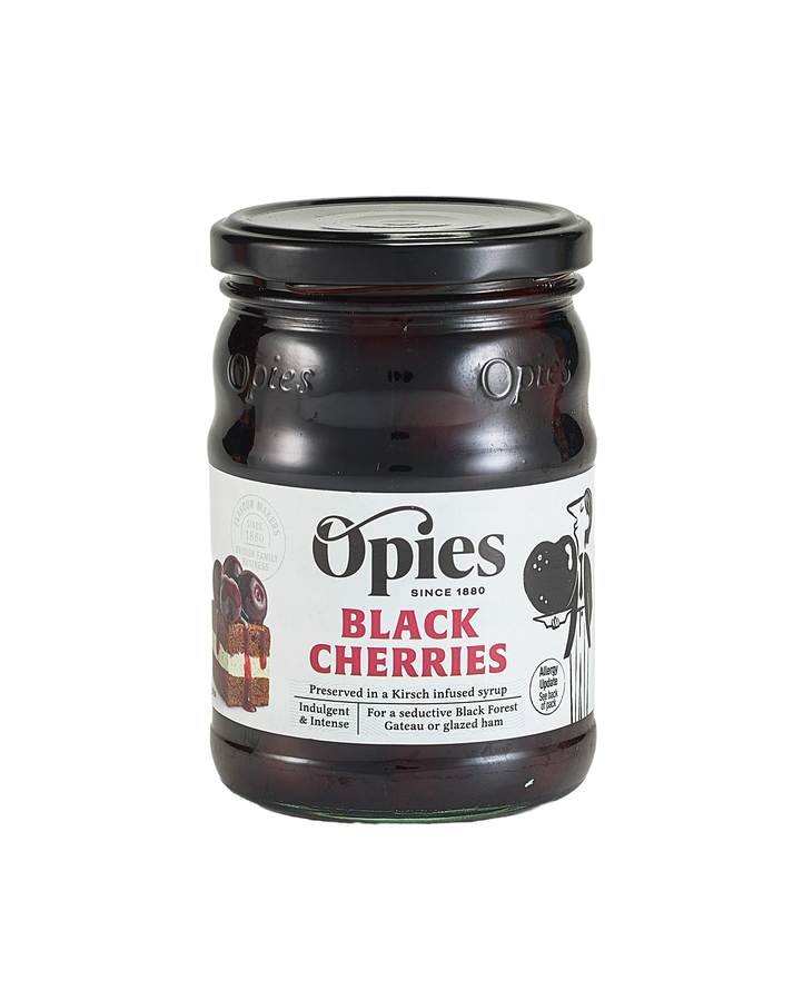 A jar of Opies Black Cherries With Kirsch, a luxurious treat featuring cherries preserved in Kirsch-infused syrup. The label displays enticing images of cherries and desserts, with text emphasizing its perfect use to adorn cakes or elevate indulgent desserts such as Black Forest gateau or glazed ham. The jar boasts a black lid and a transparent glass body, showcasing the rich contents within.