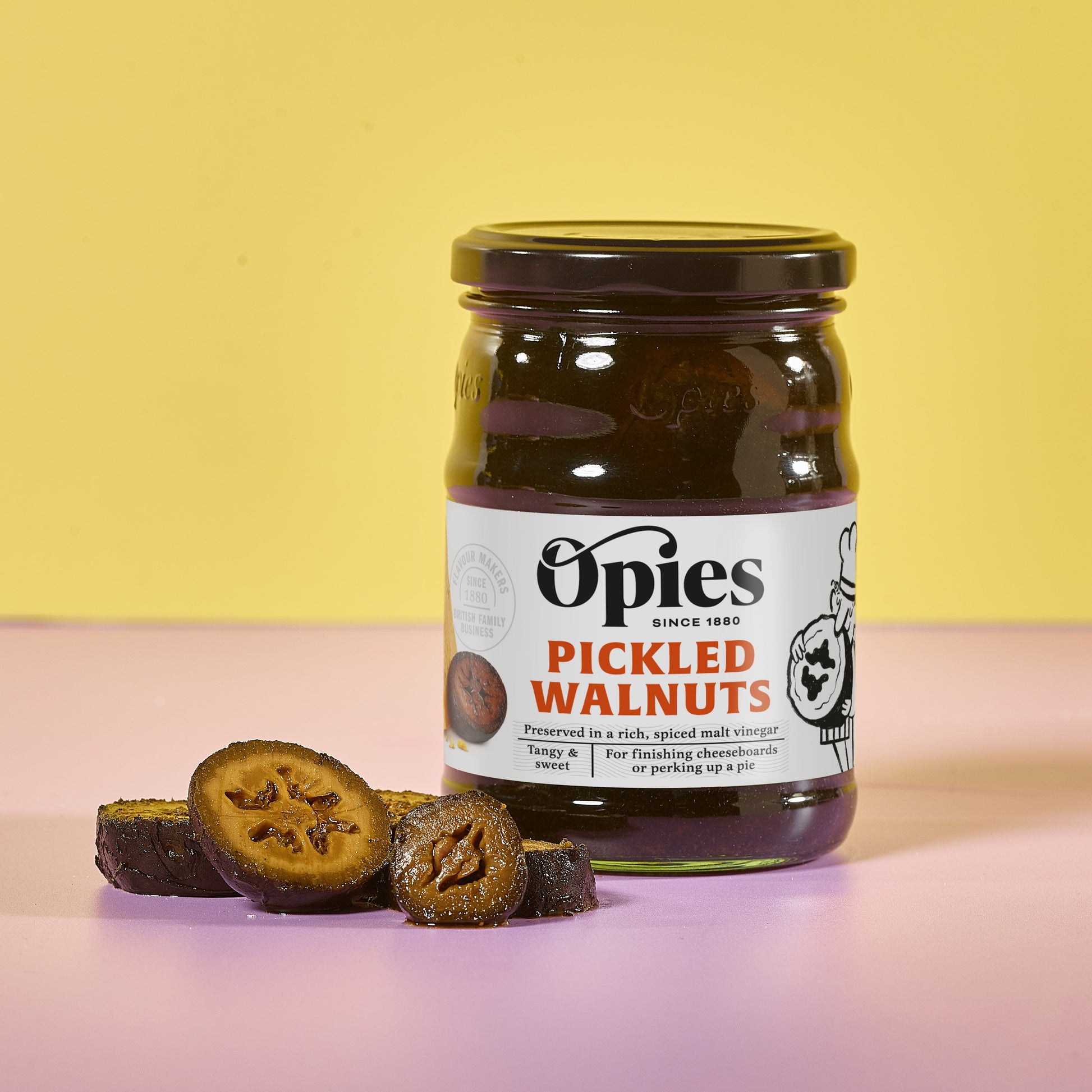 A jar of Opies Pickled Walnuts by Opies is displayed against a yellow and pink background. The jar's label reads, "Preserved in rich, spiced malt vinegar, tangy & sweet." Slices of pickled walnuts are arranged in front to reveal their interior texture—ideal for enhancing a beef casserole.