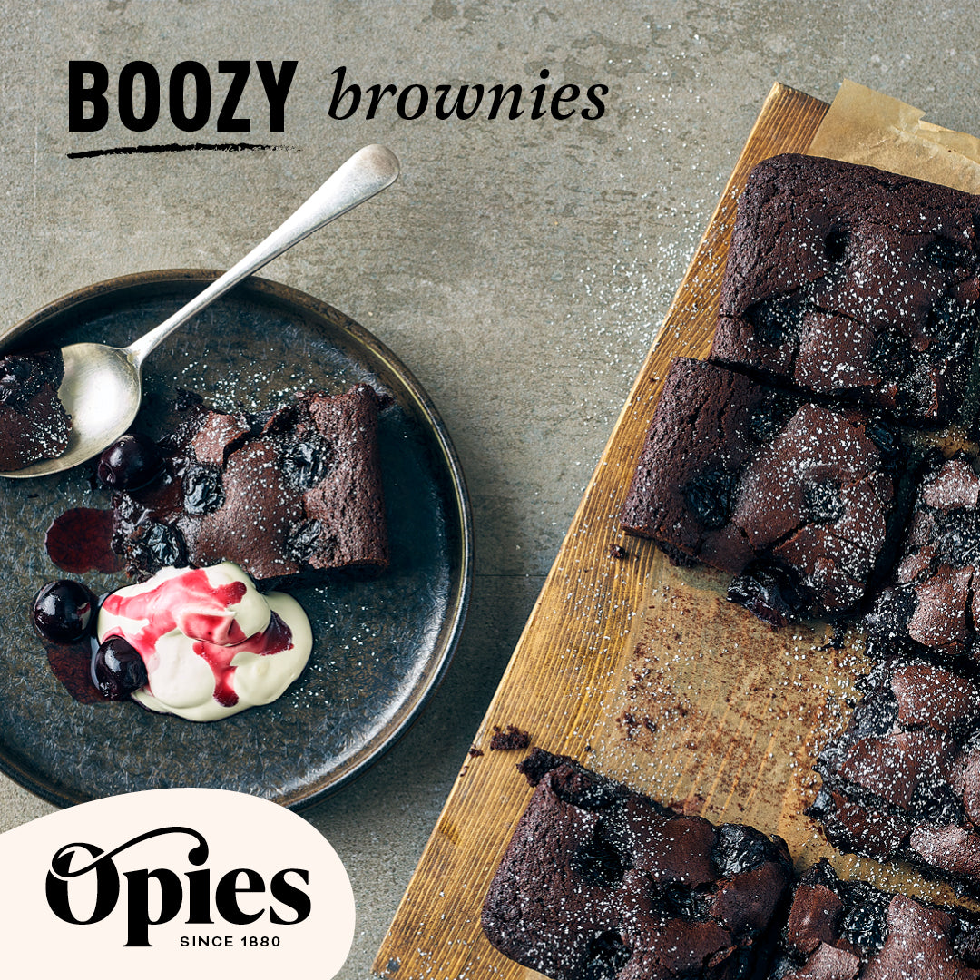 A wooden board holds several chocolate brownies with a dusting of powdered sugar. Nearby, a plate features a serving of the brownies crowned with cream and luxurious Opies Black Cherries With Kirsch. The word "BOOZY brownies" is written in black, and the "Opies since 1880" logo is displayed at the bottom left.