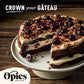 A decadent chocolate cake adorned with luxurious Opies Black Cherries With Kirsch and chocolate shavings is displayed on a white cake stand. The cake has a slice missing, revealing layers of rich chocolate sponge and cream. Text at the top reads "Crown your Gâteau," and the Opies logo is at the bottom left.