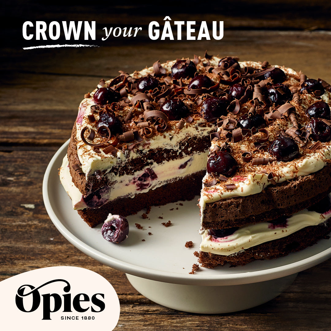 A decadent chocolate cake adorned with luxurious Opies Black Cherries With Kirsch and chocolate shavings is displayed on a white cake stand. The cake has a slice missing, revealing layers of rich chocolate sponge and cream. Text at the top reads "Crown your Gâteau," and the Opies logo is at the bottom left.