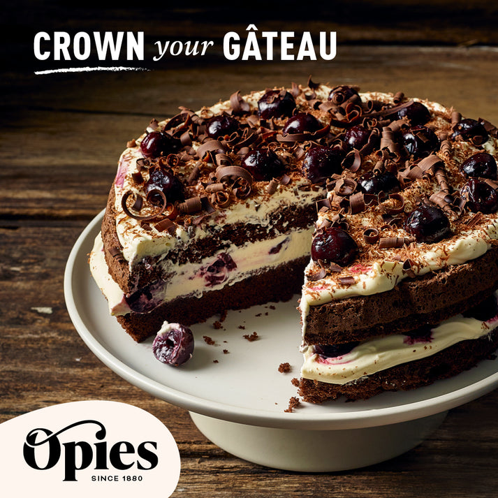 A decadent chocolate cake adorned with luxurious Opies Black Cherries With Kirsch and chocolate shavings is displayed on a white cake stand. The cake has a slice missing, revealing layers of rich chocolate sponge and cream. Text at the top reads "Crown your Gâteau," and the Opies logo is at the bottom left.