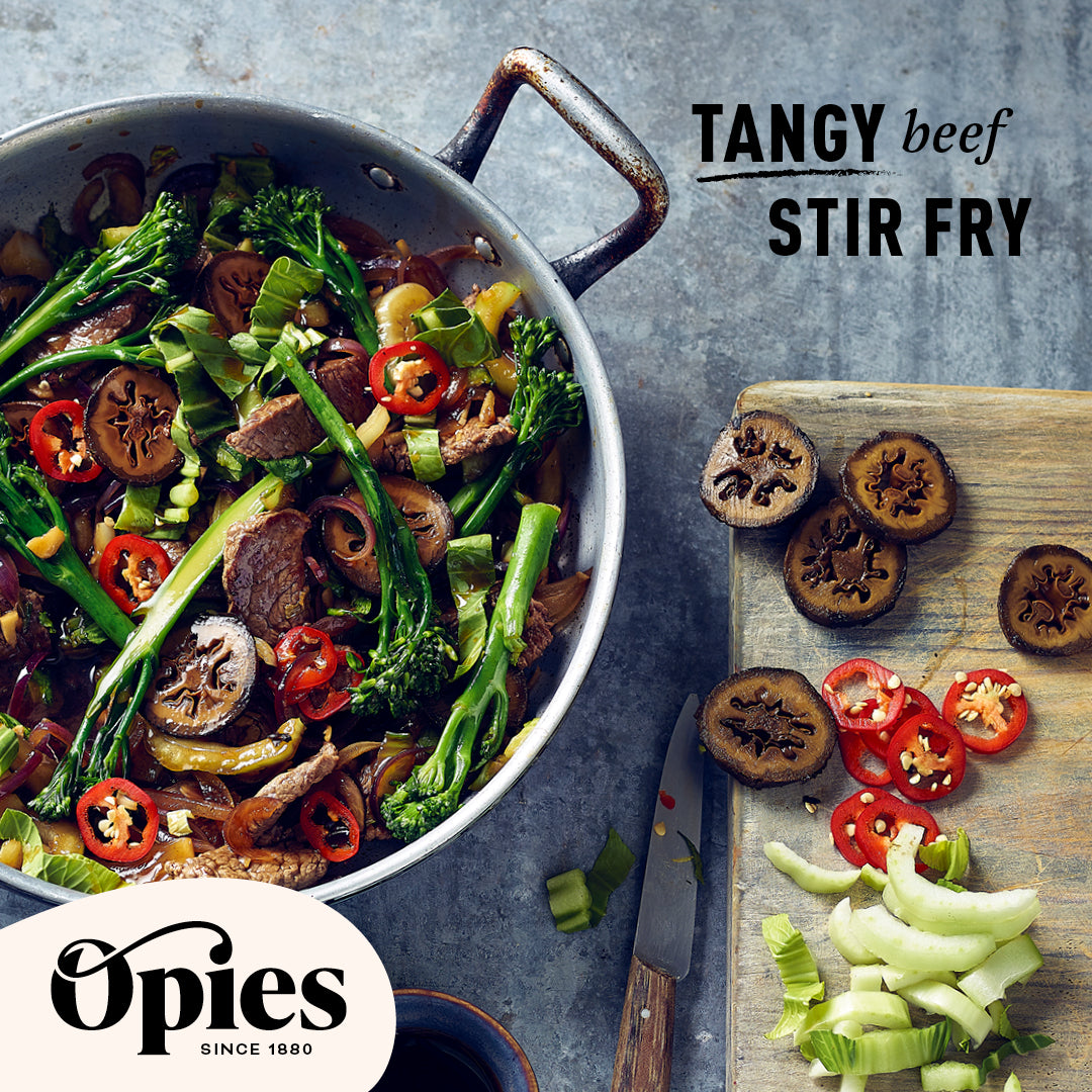 A skillet filled with a colorful, tangy beef stir fry featuring broccoli, red chili slices, beef strips, and Opies Pickled Walnuts. Next to the skillet, sliced vegetables and knives are visible. The text "TANGY beef STIR FRY" is displayed prominently. The Opies logo is at the bottom left.