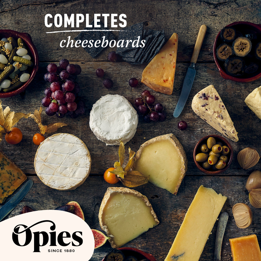 A variety of cheeses are artfully arranged on a wooden table, accompanied by grapes, olives, Opies Pickled Walnuts, and a knife. The text reads "Completes Cheeseboards" and features the Opies logo with the caption "Opies Since 1880.