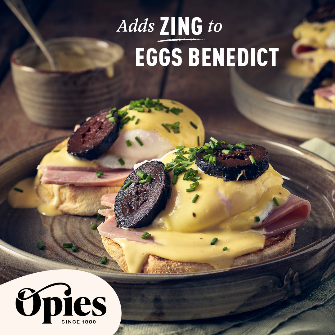 A close-up of Eggs Benedict topped with circular slices of black pudding and garnished with chives. The dish is served on a rustic plate, with a bowl of Opies Pickled Walnuts in the background. Text reads, "Adds ZING to EGGS BENEDICT." Opies logo and “Since 1880” are displayed.
